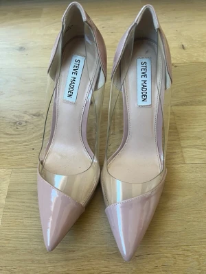 Steve Madden pumps med transparent detalj - Säljer ett par snygga pumps från Steve Madden i ljusrosa lack med spetsig tå och transparenta sidor i plast. Klackarna har en modern och stilren look som verkligen sticker ut. Perfekta för dig som vill ha något trendigt och unikt i garderoben.