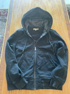 Burberry kofta - Snygg svart kofta från Burberry med dragkedja framtill och två fickor. Huvan har ett klassiskt rutmönstrat foder i grått och rött samt dragsko. Perfekt för en chill och stilren look. Materialet känns mjukt och bekvämt.