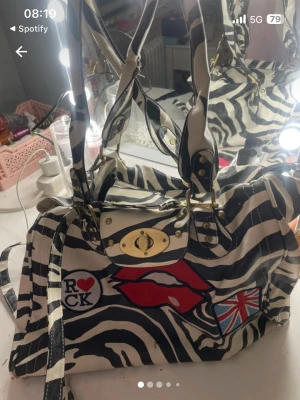 Zebramönstrad handväska med patches - Cool handväska med zebramönster i svart och vitt, dekorerad med patches som röda läppar, brittiska flaggan och en 'I ♥ Rock'-badge. Väskan har guldfärgade metalldetaljer, dubbla handtag och axelrem. Perfekt för dig som gillar statement-accessoarer.