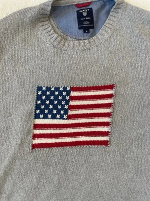 Grå stickad tröja från Gant - Snygg grå stickad tröja från Gant med amerikansk flagga broderad på bröstet. Tröjan har rund halsringning, ribbade muddar och är tillverkad i mjukt material. Perfekt för dig som gillar klassisk college-stil.