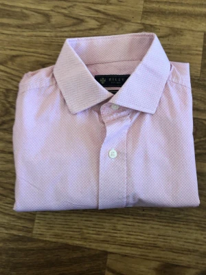 Ljusrosa slim fit skjorta från Riley - Snygg ljusrosa skjorta från Riley i slim fit-modell. Skjortan har ett diskret mönster i vitt, klassisk krage och vita knappar framtill samt vid ärmsluten. Perfekt för dig som gillar stilrena detaljer och en smalare passform.