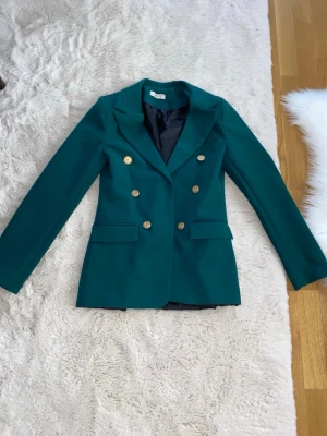 Grön dubbelknäppt kavaj med guldknappar - Stilren grön kavaj med dubbelknäppning och sex dekorativa guldknappar framtill. Klassisk krage och två fickor med lock. Svart innerfoder för extra komfort. Perfekt för att lyfta din outfit med en elegant touch.