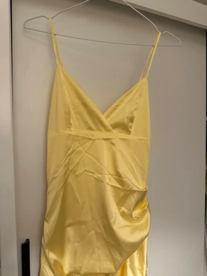 Gul långklänning med volang - Säljer en snygg gul långklänning i satinliknande material med smala axelband och v-ringad omlott upptill. Klänningen har volangdetaljer längs nederdelen som ger en extra touch. Perfekt för dig som vill sticka ut med en färgglad och elegant look. Ny utan prislapp 