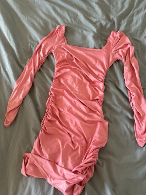 Rosa långärmad omlottklänning - Trendig rosa kortklänning med långa ärmar och snygga rynkade detaljer längs sidorna och framtill. Klänningen har en fyrkantig ringning och är tillverkad i ett mjukt, stretchigt material som ger en smickrande passform.