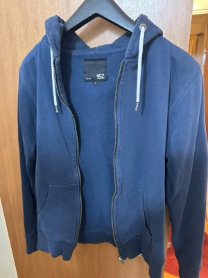 Mörkblå hoodie med dragkedja 157 - Mörkblå hoodie från 157 med dragkedja framtill, två fickor och justerbar huva med snören. Tillverkad i mjukt sweatshirtmaterial, perfekt för en avslappnad stil. Klassisk passform och ribbade muddar vid ärmar och nederkant.