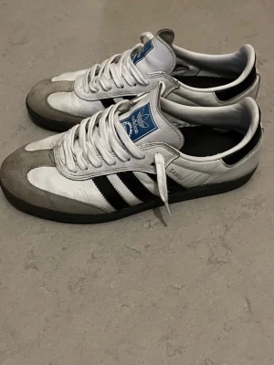 Adidas Samba vita och svarta sneakers - Klassiska Adidas Samba sneakers i vitt läder med grå mockadetaljer och svarta ränder på sidorna. Skorna har blå Adidas-logga på plösen och svart sula. Perfekta för en sportig och stilren look.