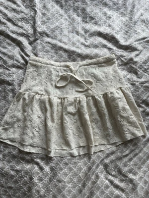 Vit kortkjol med spets och volang - Söt vit kortkjol med broderad spets och volangkant. Kjolen har en dekorativ knytning i midjan och är perfekt för sommarens alla tillfällen. Tillverkad i ett lätt och luftigt material som ger en romantisk känsla.