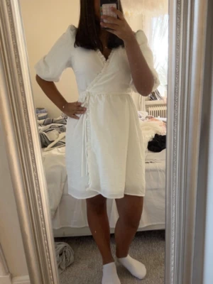 Vit omlottklänning med volang - Säljer en söt vit omlottklänning med volangdetaljer längs kanten och knyt i midjan. Klänningen har korta puffärmar och är tillverkad i ett lätt, strukturerat tyg som känns luftigt. Perfekt för varma dagar och ger en fräsch look.