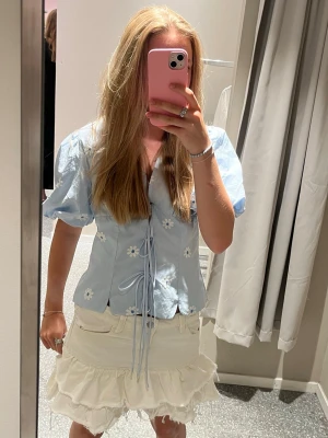 Blå blus med puffärm från missay  - Supersöt ljusblå blus från missay med puffärmar och broderade vita blommor. Blusen har knytband framtill och v-ringning som ger en feminin look. Tillverkad i ett lätt bomullstyg, perfekt för sommardagar.