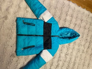 Fila. Blå och svart pufferjacka med huva - Snygg pufferjacka i blått med svarta och vita detaljer. Jackan har två dragkedjefickor framtill, huva och breda färgblock. Perfekt för kalla vinterdagar och ger en sportig vibe. Materialet är glansigt syntet och jackan har en rejäl dragkedja framtill.