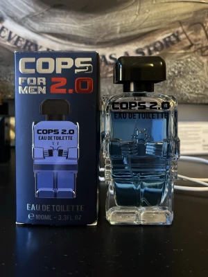 COPS 2.0 Eau de Toilette för män - COPS 2.0 Eau de Toilette för män i en unik flaska formad som en polisfigur. Flaskan är genomskinlig med blått innehåll och svart lock. Förpackningen är blå med silver- och rödfärgad text. Volymen är 100 ml.