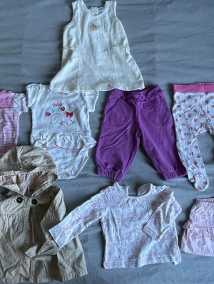 Sött babyklädpaket i pastellfärger - Flera babyplagg i mjuka material, bl.a. vit klänning, rosa och vita bodys med tryck, lila byxor, mönstrade byxor med fot, beige jacka med huva och vit långärmad tröja med rosa mönster. Fina detaljer som volanger, tryck med djur och jordgubbar samt knappar.
