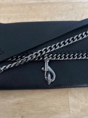 Rebecca Minkoff Svart axelremsväska med kedja - Rebecca Minkoff. Stilren svart kuvertväska i skinnimitation med silverfärgad kedja och metallspänne framtill. Väskan har en enkel, rektangulär form och detaljer i metall som ger en edgy vibe. Perfekt för dig som gillar minimalistisk design med en twist.