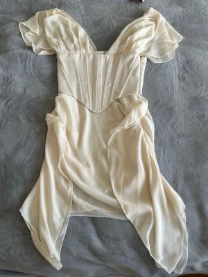 Champagnefärgad omlottklänning - Romantisk kortklänning i champagnefärgad, lätt och flytande polyester med omlottdetaljer och offshoulder-ärmar. Klänningen har markerad midja och volangdetaljer som ger en drömmig siluett. Perfekt för sommarens alla tillfällen.