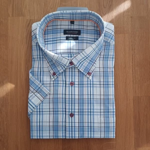 Blårutig kortärmad skjorta - Snygg blårutig skjorta från Shirt of Sweden med korta ärmar och röda knappar. Skjortan har klassisk krage och är gjord i mjuk bomull, perfekt för varma dagar. 
