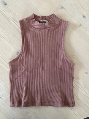 Ribbad ärmlös topp från 157 - Säljer en ribbad, croppad topp från 157 i en snygg dammrosa färg. Toppen är ärmlös med hög hals och har en tight passform som passar perfekt till jeans eller kjol. Köparen står för frakt.