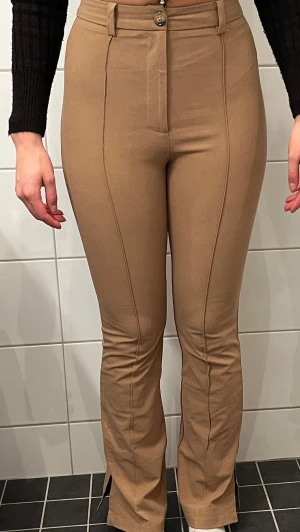 Beiga utsvängda byxor med söm - Säljer ett par beiga byxor med utsvängda ben och markerad söm framtill. Byxorna har hög midja och är tillverkade i ett stretchigt material som sitter tajt upptill och släpper nedtill. Perfekta för dig som gillar en trendig och bekväm look.