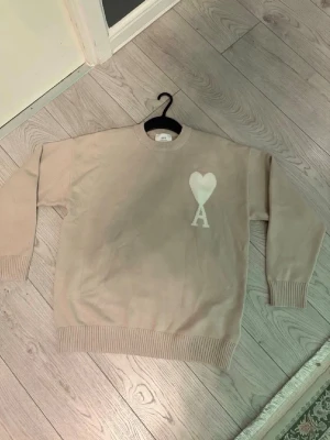 Beige stickad tröja från Ami Paris - Säljer en beige stickad tröja från Ami Paris med deras ikoniska vita hjärtat.  Perfekt för dig som gillar stilrena plagg med en twist. Helt ny kommer med tagg. Pris kan diskuteras!
