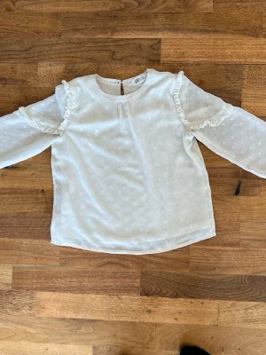 Vit blus med volangdetaljer - Söt vit blus med diskret prickstruktur och volangdetaljer längs axlarna. Blusen har rund halsringning, knapp i nacken och långa, transparenta ärmar med knapp vid ärmslut. Perfekt för en trendig och feminin look.