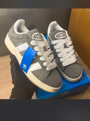Adidas Campus grå sneakers - Snygga Adidas Campus sneakers i grå mocka med vita detaljer och breda vita skosnören. Klassisk låg modell med gummisula och ikoniska tre ränder på sidan. Perfekta för en clean och stilren look.