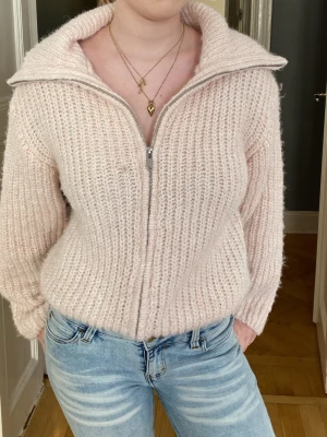 Ljusrosa stickad ziptröja - Mysig ljusrosa stickad tröja med dragkedja framtill och stor krage. Tröjan har en fluffig struktur och är ribbstickad, vilket ger en avslappnad och trendig look. Passar perfekt till jeans och är ett måste i garderoben för kyliga dagar. Har en liten foundation fläck framtill. 