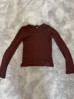 Brun långärmad topp från H&M - Mörkbrun långärmad topp från H&M med enkel och stilren design. Tillverkad i mjukt material som sitter tight mot kroppen. Perfekt basplagg att matcha med olika stilar. Den är för liten för mig på bilden.