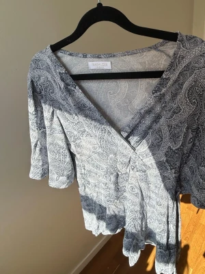 Grå omlottopp från Rabalder - Supersnygg grå omlottopp från Rabalder med paisleymönster. Toppen har v-ringning, korta ärmar och knyt i midjan för en skön passform. Perfekt till jeans eller kjol för en trendig look.