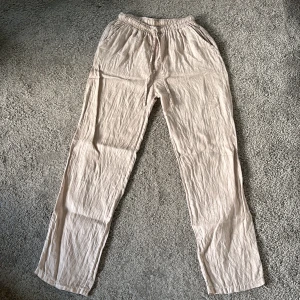 Beiga linnebyxor från Design by Si - Säljer ett par beiga linnebyxor från Design by Si med resår i midjan och raka ben. Perfekta för varma dagar och har en avslappnad passform. Byxorna har fickor i sidorna och är tillverkade i ett lätt och luftigt material.