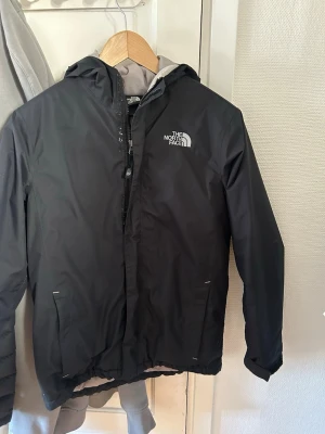 The North Face jacks - Snygg svart regnjacka från The North Face med huva och dragkedja framtill. Jackan har logga på bröstet och ryggen, samt praktiska fickor. Perfekt för blöta dagar och enkel att matcha med allt. Lätt och smidig design. Det är storlek L i junior men sitter som en XS/S