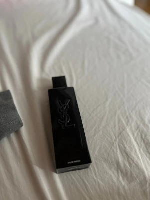 YSL svart parfymflaska - Säljer en elegant parfym från Yves Saint Laurent i en svart, rektangulär flaska med YSL-loggan i relief på framsidan. Flaskan har en modern och stilren design som känns lyxig och passar perfekt på parfymhyllan.