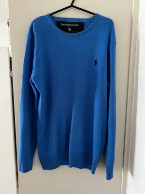 Blå stickad tröja Polo Ralph Lauren - Snygg blå stickad tröja från Polo Ralph Lauren med rund halsringning och diskret broderad logga på bröstet. Tröjan har långa ärmar och ribbade muddar vid ärmslut och nederkant. Perfekt för lager på lager och enkel att matcha.