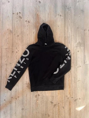 Svart Kenzo hoodie med tryck - Snygg svart hoodie från Kenzo med stort vitt tryck på båda ärmarna där det står 'KENZO PARIS'. Klassisk huva och ribbade muddar. Perfekt för dig som gillar streetwear och vill ha något stilrent men ändå med attityd.
