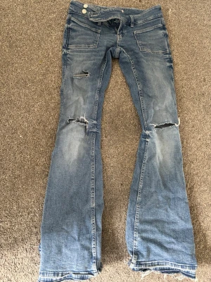 Blå slitna bootcut jeans - Säljer ett par blå bootcut jeans med slitningar och hål på benen för en trendig look. Jeansen har klassiska fickor fram och bak samt en knappgylf. Tillverkade i denim med en snygg tvättad finish och råa kanter vid benslut. Inga fel med dom bara att hålet har sprättas upp lite och dom e uppsprättade längst ner pris kan diskuteras❣️❣️