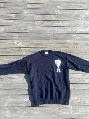 Mörkblå stickad tröja från Ami - Säljer en mörkblå stickad tröja från Ami med rund halsringning och ett vitt hjärta med bokstaven 'A' broderat på bröstet. Tröjan har långa ärmar och ribbade muddar vid ärmslut och nederkant. Perfekt för dig som gillar stilrena detaljer.