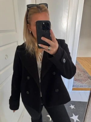 Svart skepparkavaj från VILA - Stilren svart skepparkavaj från VILA med dubbelknäppning och breda slag. Klassisk kort modell och mjukt ull-liknande material. Perfekt för dig som vill ha en tidlös och trendig jacka till hösten eller våren!!