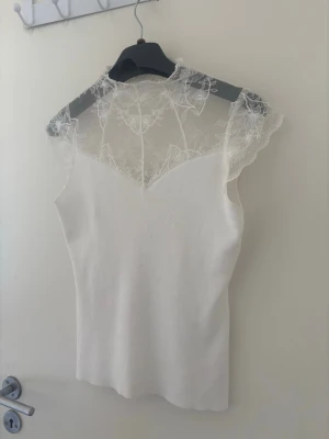 Vit topp med spetsdetaljer - Säljer en elegant vit topp med transparent spets upptill och vid axlarna. Toppen har en figurnära passform och är ärmlös med vacker spets som ger en romantisk känsla. Perfekt att styla med jeans eller kjol för en trendig look.