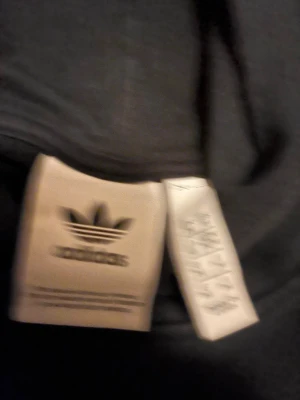 Svart hoodie från Adidas med tryck - Säljer en svart hoodie från Adidas med färgglatt tryck på bröstet och klassisk känguruficka. Hoodien har huva med dragsko och är tillverkad i mjukt material, perfekt för en avslappnad streetwear-look.