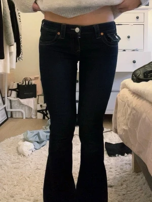 true religion jeans - Säljer ett par mörkblå bootcut jeans från True Religion med klassiska kontrastsömmar och dekorativa ficklock bak. Jeansen har låg midja och utsvängda ben, tillverkade i stretchigt denimtyg för skön passform. midje måttet är 38 cm