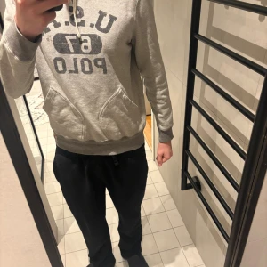 Grå hoodie från U.S. Polo Assn - Säljer en grå hoodie från U.S. Polo Assn med stor logga och text på bröstet. Hoodien har huva med dragsko, magficka och ribbade muddar vid ärmslut och nederkant. Perfekt för en avslappnad och sportig stil.