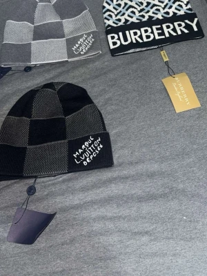 Burberry och Louis Vuitton mössor - Säljer tre trendiga mössor: en svart och grå rutmönstrad Louis Vuitton, en ljusgrå och vit rutmönstrad Louis Vuitton samt en svartvit Burberry med grafiskt mönster och stor logga. Alla mössor är stickade och har tydliga logotyper framtill.