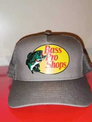 Grå Bass Pro Shops keps - Snygg grå keps från Bass Pro Shops med klassisk gul och röd logga framtill och en stor fiskgrafik. Kepsen har mesh på sidorna och justerbar snapback baktill. Perfekt för dig som gillar att fiska eller bara vill ha en keps som funkar till allt!