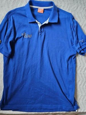 Blå pikétröja från Puma - Blå pikétröja från Puma med vit knappslå och broderad text '24h GOLF' i guld på bröstet. Klassisk krage och korta ärmar. Perfekt för dig som gillar sportig stil och vill ha något bekvämt och snyggt.