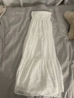 Vit långkjol med volanger Gina Tricot - Lång, vit kjol från Gina Tricot med smock i midjan och diskret strukturmönster i tyget. Kjolen har flera volanger och är tillverkad i ett lätt och luftigt material, perfekt för varma dagar. Använd en gång 