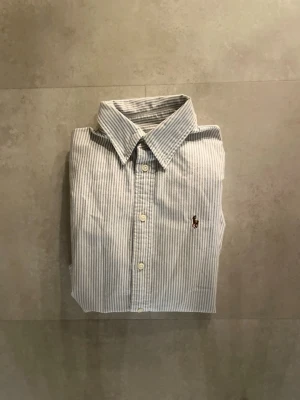 Randig skjorta från Polo Ralph Lauren - Klassisk ljusblå och vit randig skjorta från Polo Ralph Lauren i bomull. Custom fit-modell med knappstängning framtill och broderad logga på bröstet. Perfekt för en stilren och tidlös look.