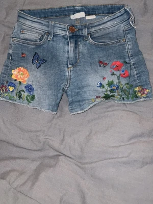 Blå jeansshorts med blommigt broderi - Säljer ett par blå jeansshorts med färgglatt broderi av blommor, fjärilar och nyckelpigor framtill. Midjemått: Ca 30 cm Grenen: 20 cm. Är storlek XXS-XS