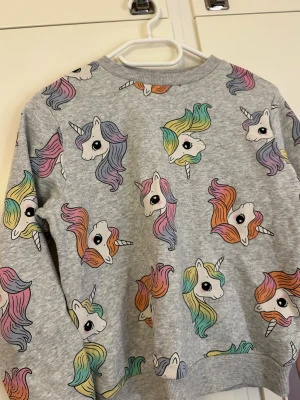 Grå sweatshirt med enhörningsmönster - Grå sweatshirt med färgglada enhörningar i pastellfärger över hela tröjan. Rund halsringning och ribbade muddar vid ärmslut och nederkant. Perfekt för dig som gillar lekfulla och unika prints.