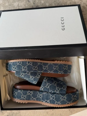 Gucci platåsandaler i denim - Snygga platåsandaler från Gucci i blå denim med det klassiska GG-mönstret. Sandalerna har breda remmar och grov sula i brunt och beige. Perfekta för dig som vill ha en trendig och lyxig look med streetvibes.