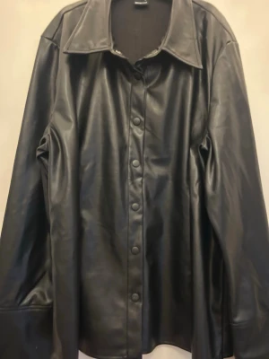 Svart skinnskjorta med tryckknappar - Stilren svart skinnskjorta  med klassisk krage och tryckknappar framtill. Skjortan har raka ärmar och en något oversized passform som ger en cool look. Perfekt att slänga över en hoodie eller t-shirt för en edgy vibe. Storlek XL