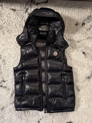 Moncler Väst - Snygg svart dunväst från Moncler med glansig finish och avtagbar luva. Västen har två dragkedjeförsedda fickor och klassisk Moncler-logga på bröstet. Perfekt för lager-på-lager och streetstyle. Materialet är dunfyllning med yttertyg i nylon. Allt från köpet på Nk såsom kvitto medföljer.
