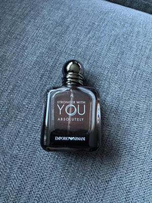 Emporio Armani Stronger With You - Emporio Armani Stronger With You Absolutely är en elegant parfym i en stilren, fyrkantig glasflaska med mörk toning och en blank, rund silverfärgad kork. Flaskan har tydlig logotyp och text framtill, vilket ger en lyxig och modern känsla.
